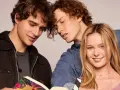 Die "Love Me, Love Me" Besetzung: Zwei junge Männer lesen ein Buch, während eine junge Frau lächelnd in die Kamera blickt.