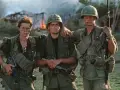 Die Besetzung von Platoon: Drei Soldaten in Uniform, mit Waffen und Ausrüstung, stehen vor brennenden Gebäuden.