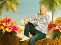 Ein Mann sitzt in einer Kokosnussschale, umgeben von tropischen Blumen und Palmen. Ein Bild, das an Filme von Claude-Oliver Rudolph erinnert.
