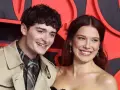 Noah Schnapp und eine Frau lächeln vor einem roten Hintergrund. Er trägt einen Trenchcoat und eine karierte Krawatte, wie man sie aus seinen Filmen kennt.