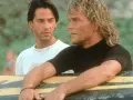 Keanu Reeves und Patrick Swayze, die Hauptdarsteller der Besetzung von Point Break, lehnen an einem Surfbrett.