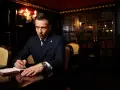 Ein Mann im Anzug schreibt in einem Notizbuch. Er könnte Teil der **Besetzung von The Lincoln Lawyer** sein, mit einem Bücherregal im Hintergrund.