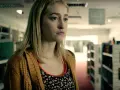 Junge Frau mit blonden Haaren und einem gelben Strickcardigan steht in einem Gang mit Bücherregalen. Ein Ausschnitt aus Gus Birneys Filme und Serien.