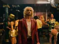 Ein Mann mit blonden Haaren und rotem Fransen-Sakko steht im Mittelpunkt, umgeben von einer bunten **Besetzung von Luden** in einem Nachtclub mit Discokugel.