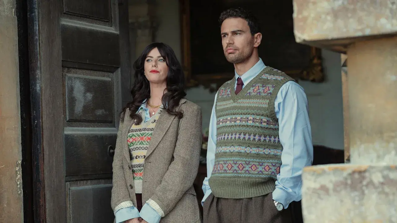 Die Besetzung von The Gentlemen: Ein Mann und eine Frau stehen vor einer Holztür, beide in Strickwesten und Tweed.