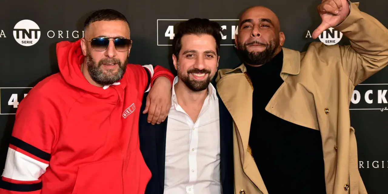 Drei Männer posieren vor einem Banner mit "4 Blocks Staffel 4". Einer trägt eine rote Nike-Jacke, ein anderer einen beigen Trenchcoat.