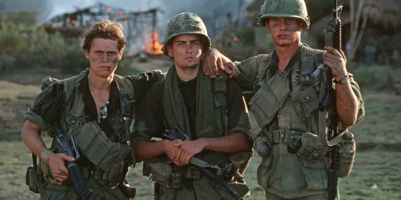 Die Besetzung von Platoon: Drei Soldaten in Uniform, mit Waffen und Ausrüstung, stehen vor brennenden Gebäuden.