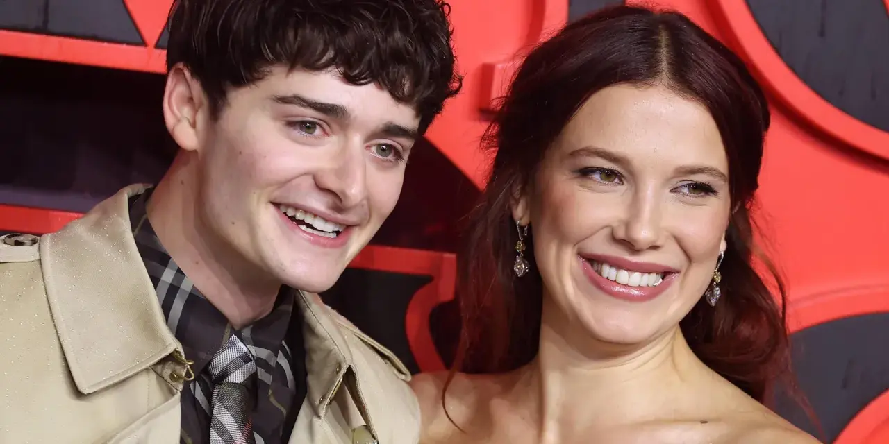 Noah Schnapp und eine Frau lächeln vor einem roten Hintergrund. Er trägt einen Trenchcoat und eine karierte Krawatte, wie man sie aus seinen Filmen kennt.