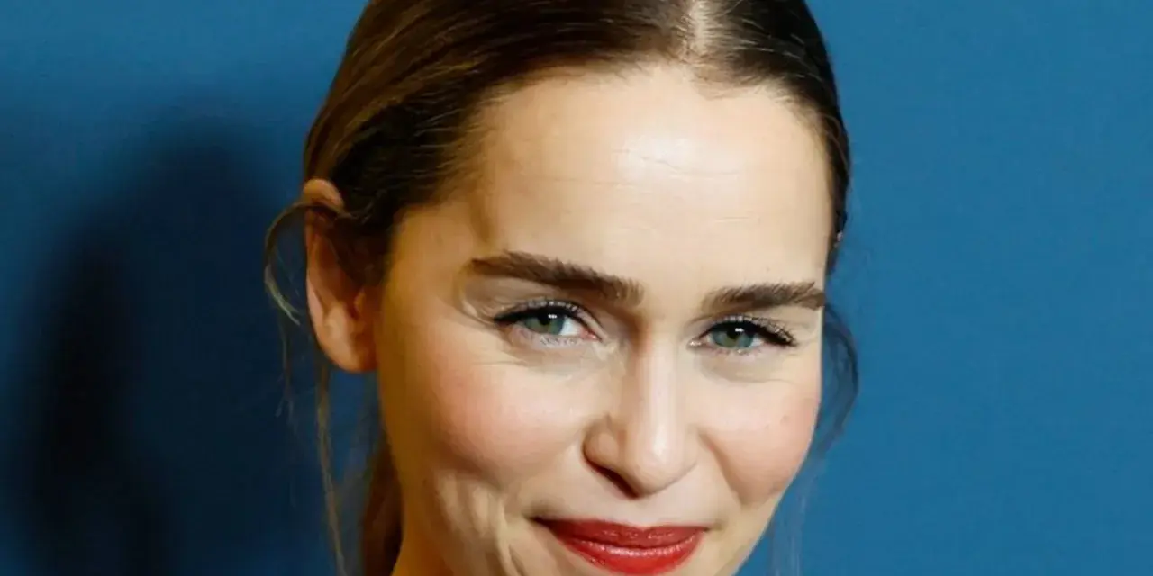 Emilia Clarke, bekannt für ihre Rollen in **filme und serien von Emilia Clarke**, lächelt charmant vor blauem Hintergrund.