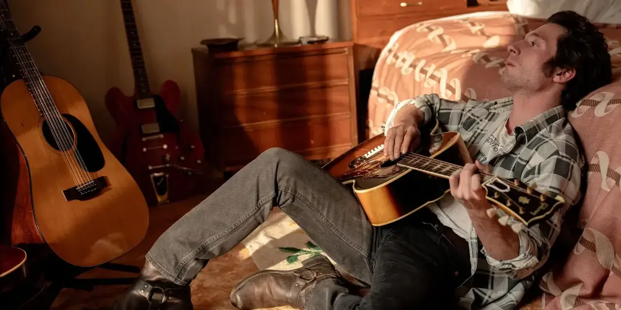 Ein Mann mit Gitarre, inspiriert von Bruce Springsteen, sitzt im Zimmer. Mehrere Gitarren stehen im Hintergrund.