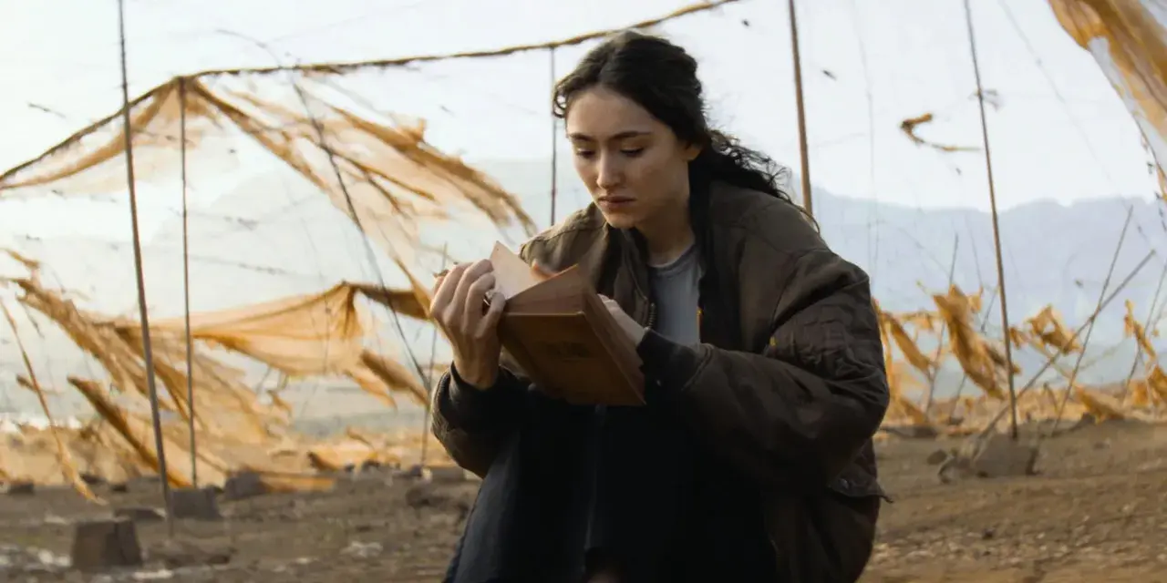 Eine junge Frau liest in einem alten Buch, umgeben von verwitterten Netzen. Ein Moment der Ruhe in der Welt von Oderbruch Staffel 3.