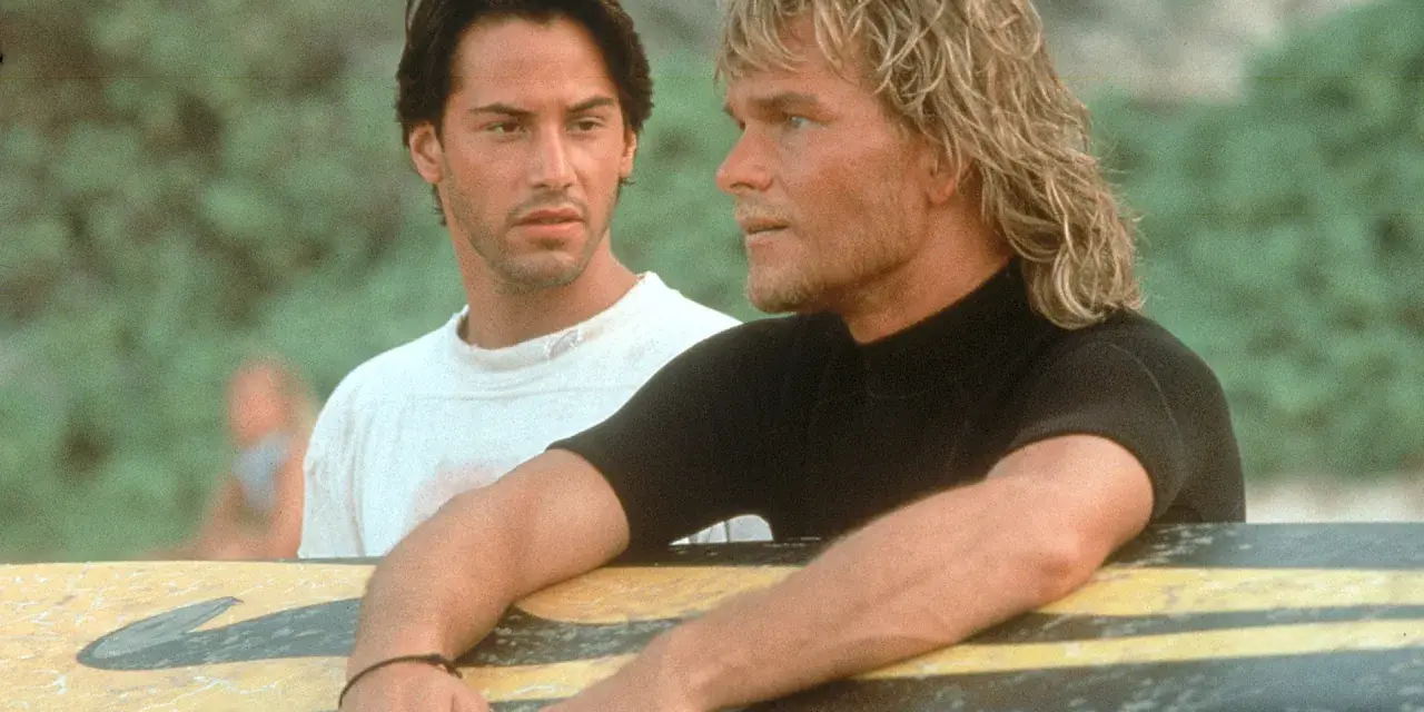 Keanu Reeves und Patrick Swayze, die Hauptdarsteller der Besetzung von Point Break, lehnen an einem Surfbrett.