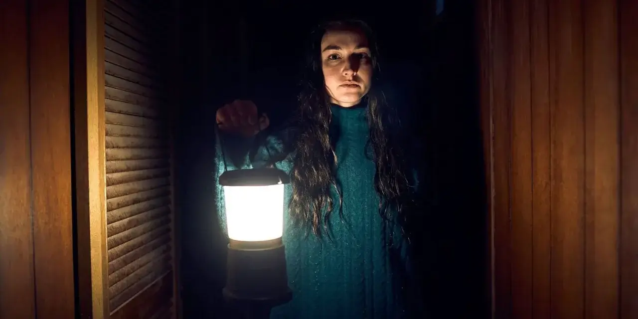 Frau mit Laterne im dunklen Flur einer Hütte. Ein Klopfen an der Kabinentür lässt sie innehalten.