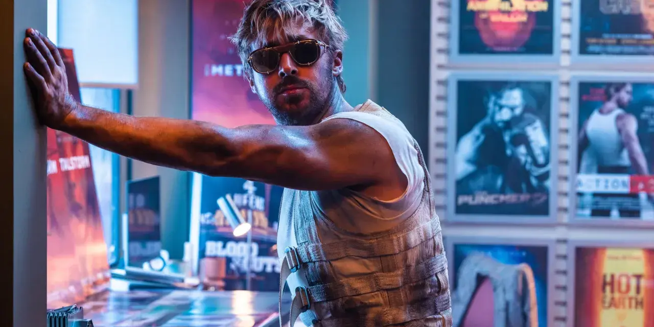 Ryan Gosling in Sonnenbrille, lehnt an einer Wand mit Postern seiner Filme.