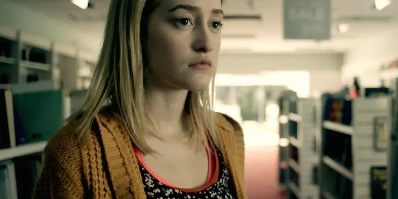 Junge Frau mit blonden Haaren und einem gelben Strickcardigan steht in einem Gang mit Bücherregalen. Ein Ausschnitt aus Gus Birneys Filme und Serien.
