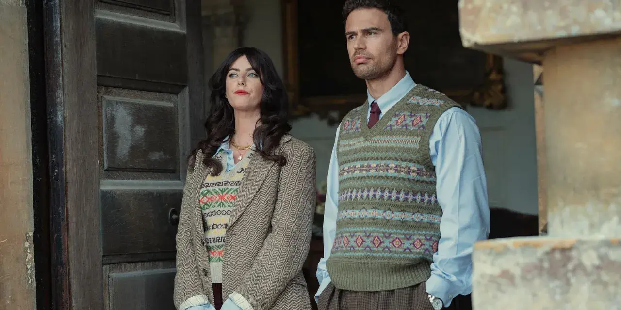 Die Besetzung von The Gentlemen: Ein Mann und eine Frau stehen vor einer Holztür, beide in Strickwesten und Tweed.