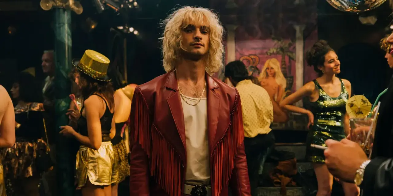 Ein Mann mit blonden Haaren und rotem Fransen-Sakko steht im Mittelpunkt, umgeben von einer bunten **Besetzung von Luden** in einem Nachtclub mit Discokugel.