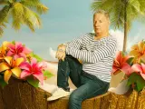 Ein Mann sitzt in einer Kokosnussschale, umgeben von tropischen Blumen und Palmen. Ein Bild, das an Filme von Claude-Oliver Rudolph erinnert.