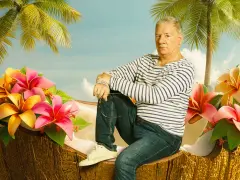 Ein Mann sitzt in einer Kokosnussschale, umgeben von tropischen Blumen und Palmen. Ein Bild, das an Filme von Claude-Oliver Rudolph erinnert.