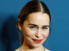 Emilia Clarke, bekannt für ihre Rollen in **filme und serien von Emilia Clarke**, lächelt charmant vor blauem Hintergrund.