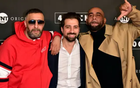Drei Männer posieren vor einem Banner mit "4 Blocks Staffel 4". Einer trägt eine rote Nike-Jacke, ein anderer einen beigen Trenchcoat.