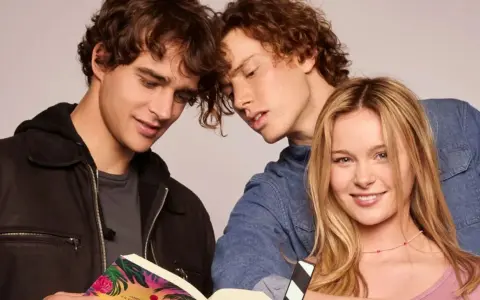 Die "Love Me, Love Me" Besetzung: Zwei junge Männer lesen ein Buch, während eine junge Frau lächelnd in die Kamera blickt.