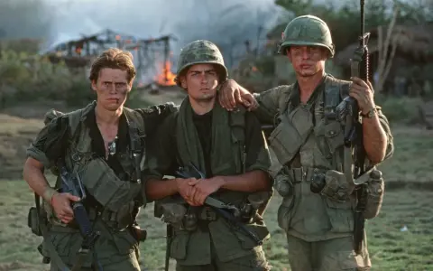 Die Besetzung von Platoon: Drei Soldaten in Uniform, mit Waffen und Ausrüstung, stehen vor brennenden Gebäuden.