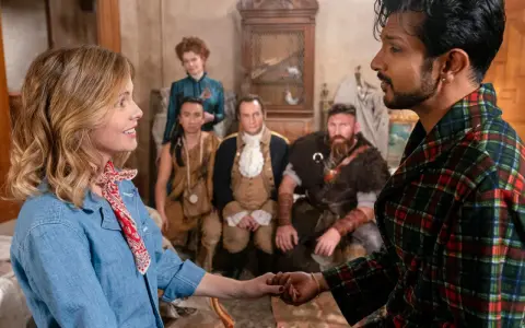 Ein Mann im karierten Bademantel hält die Hand einer Frau. Im Hintergrund sind mehrere historische Figuren zu sehen, die auf ein neues Abenteuer in "Ghosts Staffel 5" hindeuten.