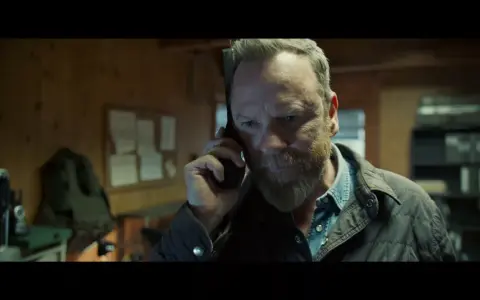 Mann mit Bart telefoniert, im Hintergrund eine Pinnwand und Regale. Besetzung von The Contractor.