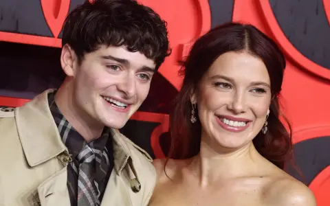 Noah Schnapp und eine Frau lächeln vor einem roten Hintergrund. Er trägt einen Trenchcoat und eine karierte Krawatte, wie man sie aus seinen Filmen kennt.