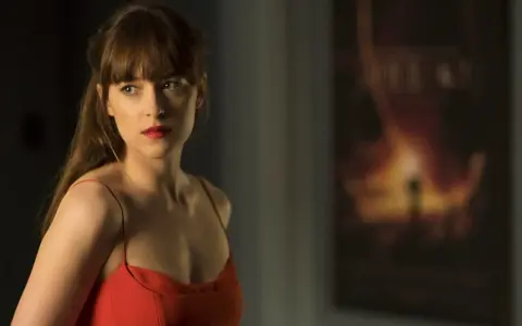 Dakota Johnson in einem roten Kleid, nachdenklich blickend. Ein Poster im Hintergrund deutet auf ihre Filme hin.