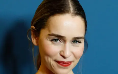 Emilia Clarke, bekannt für ihre Rollen in **filme und serien von Emilia Clarke**, lächelt charmant vor blauem Hintergrund.