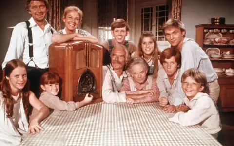 Die Besetzung von Die Waltons versammelt sich um ein Radio. Die Familie lächelt fröhlich in die Kamera.