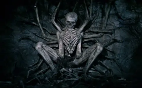 Ein skelettartiges Wesen, der leere Mann, sitzt in einer dunklen Höhle, umgeben von knochigen Gliedmaßen.