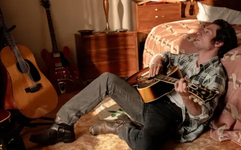 Ein Mann mit Gitarre, inspiriert von Bruce Springsteen, sitzt im Zimmer. Mehrere Gitarren stehen im Hintergrund.