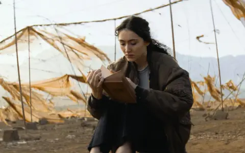 Eine junge Frau liest in einem alten Buch, umgeben von verwitterten Netzen. Ein Moment der Ruhe in der Welt von Oderbruch Staffel 3.