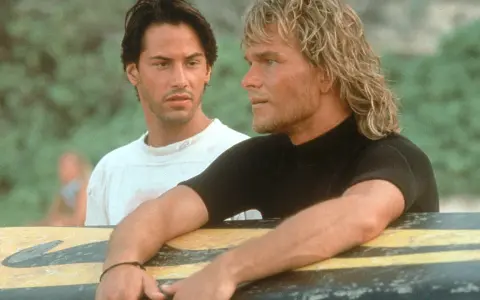 Keanu Reeves und Patrick Swayze, die Hauptdarsteller der Besetzung von Point Break, lehnen an einem Surfbrett.