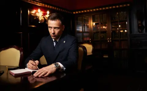 Ein Mann im Anzug schreibt in einem Notizbuch. Er könnte Teil der **Besetzung von The Lincoln Lawyer** sein, mit einem Bücherregal im Hintergrund.