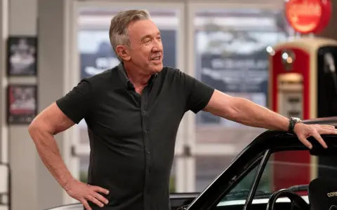 Tim Allen, bekannt für seine Filme und Serien, lehnt lässig an einem Auto. Im Hintergrund eine alte Zapfsäule.