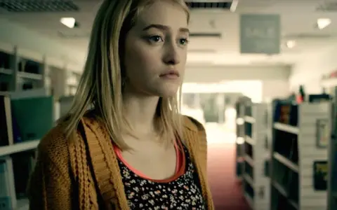 Junge Frau mit blonden Haaren und einem gelben Strickcardigan steht in einem Gang mit Bücherregalen. Ein Ausschnitt aus Gus Birneys Filme und Serien.