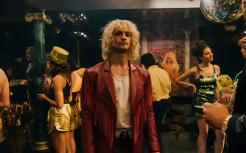 Ein Mann mit blonden Haaren und rotem Fransen-Sakko steht im Mittelpunkt, umgeben von einer bunten **Besetzung von Luden** in einem Nachtclub mit Discokugel.