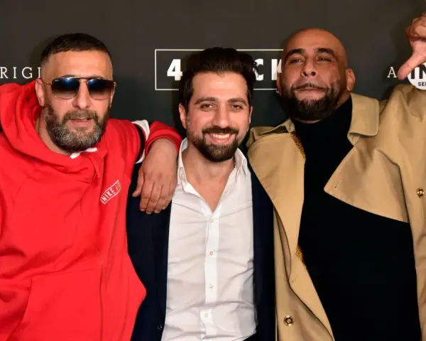 Drei Männer posieren vor einem Banner mit "4 Blocks Staffel 4". Einer trägt eine rote Nike-Jacke, ein anderer einen beigen Trenchcoat.