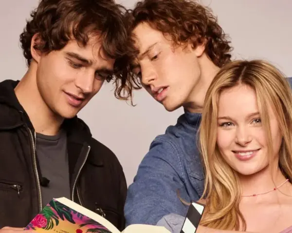 Die "Love Me, Love Me" Besetzung: Zwei junge Männer lesen ein Buch, während eine junge Frau lächelnd in die Kamera blickt.