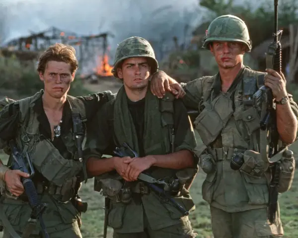 Die Besetzung von Platoon: Drei Soldaten in Uniform, mit Waffen und Ausrüstung, stehen vor brennenden Gebäuden.