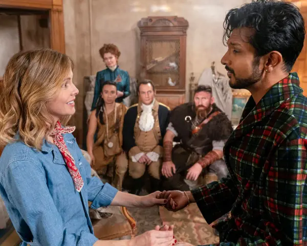 Ein Mann im karierten Bademantel hält die Hand einer Frau. Im Hintergrund sind mehrere historische Figuren zu sehen, die auf ein neues Abenteuer in "Ghosts Staffel 5" hindeuten.