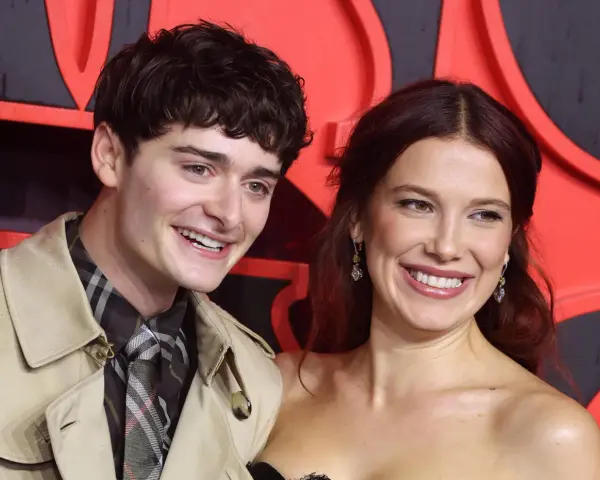 Noah Schnapp und eine Frau lächeln vor einem roten Hintergrund. Er trägt einen Trenchcoat und eine karierte Krawatte, wie man sie aus seinen Filmen kennt.