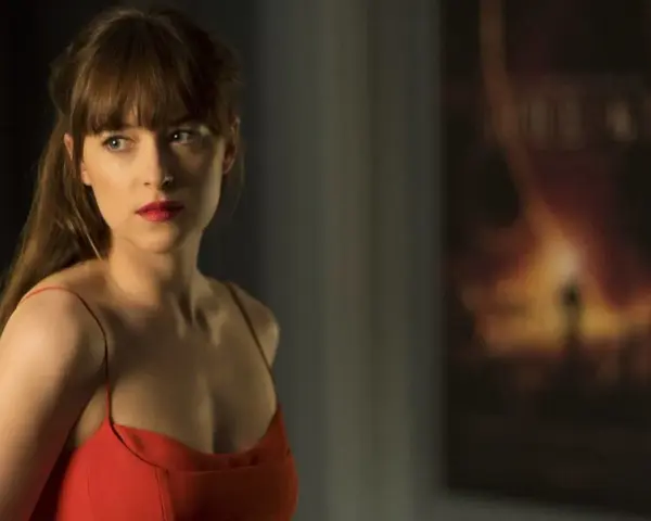 Dakota Johnson in einem roten Kleid, nachdenklich blickend. Ein Poster im Hintergrund deutet auf ihre Filme hin.