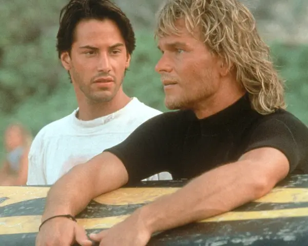 Keanu Reeves und Patrick Swayze, die Hauptdarsteller der Besetzung von Point Break, lehnen an einem Surfbrett.