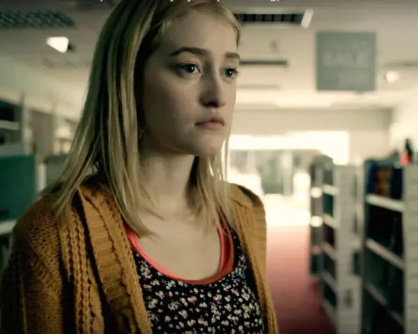 Junge Frau mit blonden Haaren und einem gelben Strickcardigan steht in einem Gang mit Bücherregalen. Ein Ausschnitt aus Gus Birneys Filme und Serien.