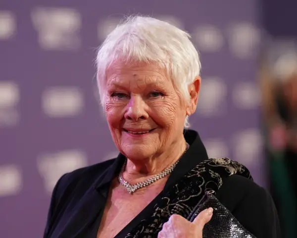 Dame Judi Dench, eine Ikone der Filme von Judi Dench, lächelt auf dem roten Teppich. Sie trägt eine schwarze Jacke, eine Diamantkette und hält eine glitzernde Clutch.